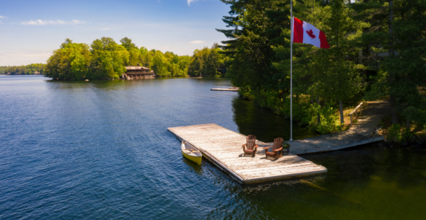 An image of a serene and still lake on a beautiful sunny day. The image shows a dock with a canoe tethered to the edge. A Canadian flag is proudly displayed. Across the lake a neighbour’s cottage can be seen.